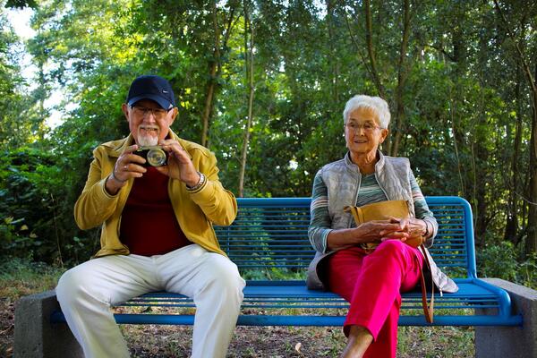 Zwei Senioren (Mann und Frau) sitzen auf Parkbank. Er hat eine Kamera in der Hand.